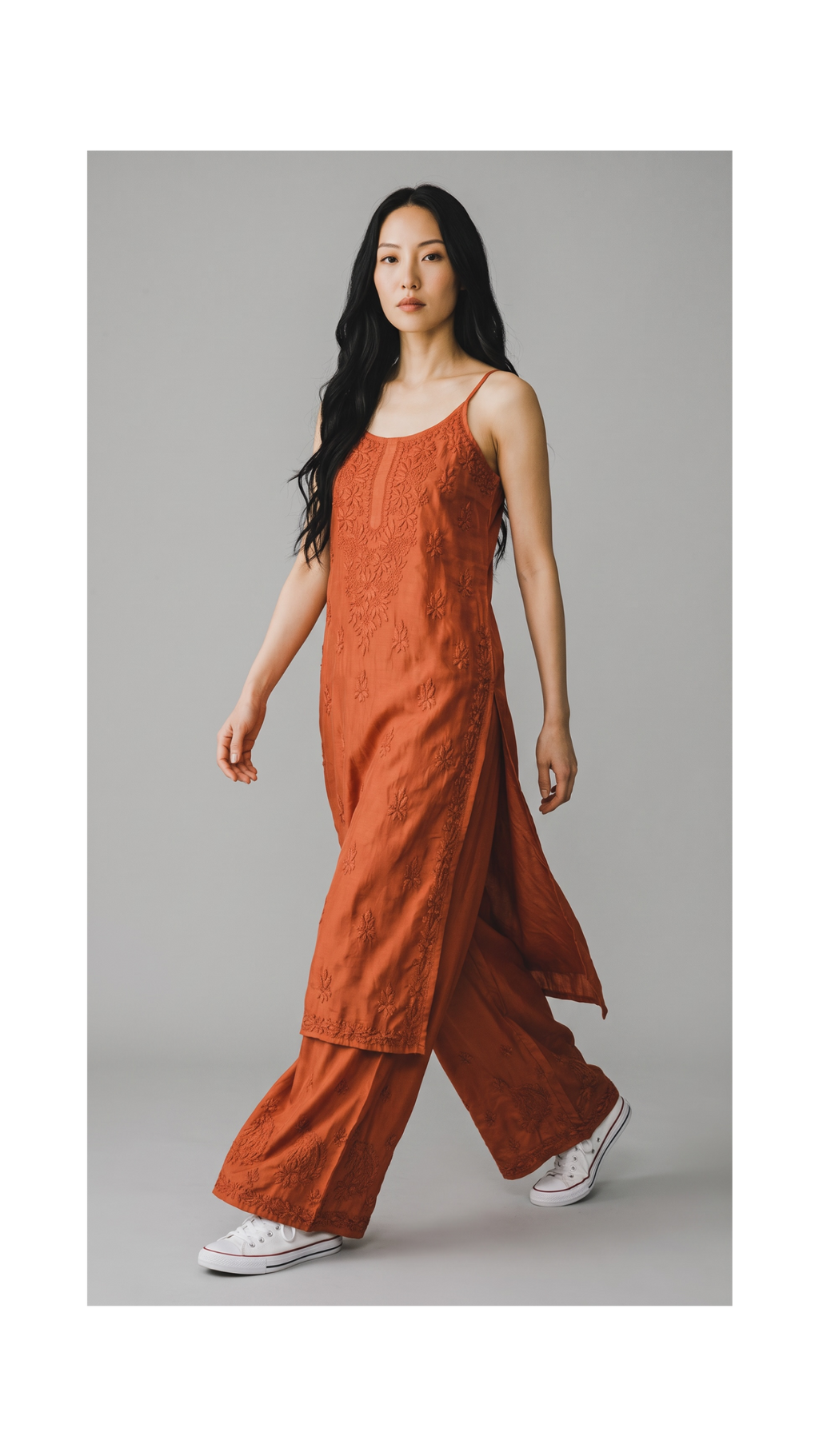 A woman modeling a rust-orange sleeveless maxi dress with wide-leg pants, featuring floral embroidery and a draped back detail, paired with white sneakers against a solid gray studio background.